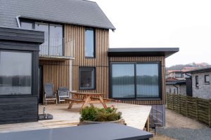 WoodUpp-fasade med tynne, vertikale trelameller i naturlig farget tre og WoodUpp-terrasse med lysbehandlede terrassebord danner husets ytre, med mørk, smal innramming rundt vinduer. Bildet viser også store skyvedører i sort ramme, en trepiknikbenk og sammenfoldede utestoler på terrasse ved vannspeil.