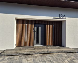 Bildet viser en inngang med mørk grå dør, omgitt av WoodUpp vertikale trelameller som fasadepanel på hver side, to smale innfelte lyslister og et metallhusnummer '13A'. Foran ligger brede betong- eller keramiske trinn og belegningsstein i gråtoner. Treverket har varm, naturlig farge og gir et moderne, minimalistisk uttrykk.