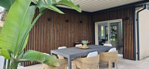 Uteoverbygd terrasse med spisebord og kurvstoler foran en vegg dekket av WoodUpp vertikale trelameller — mørke, slanke lameller montert med svarte rammer som skaper slissede paneler. Bildet viser også en plante i forgrunnen, dekorativt fat på bordet og skyvedør inn til huset.