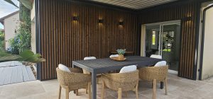 Overbygd uteplass med WoodUpp vertikal spilevegg i mørkt tre og svart ramme, og WoodUpp grå terrassebord til venstre. Mørkt spisebord med seks kurvstoler og hvite puter, kurvfat med plante på bordet og fire vegglamper som kaster en varm, koselig glød.