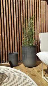 WoodUpp: bilde av vertikale trelameller i lyst, varme naturtoner med sorte mellomrom, bak to grå WoodUpp plantepotter med keramisk utseende — en stor med høyt prydgress og en mindre tom — står på tredekke, med del av hvit hagestol og rund nettingbord i forgrunnen.