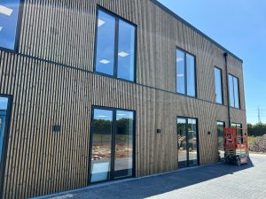 Bygning kledd med WoodUpp trelameller og fasadesystem: vertikale, smale, mørkebehandlede lameller montert med jevne avstander som gir markante skyggespill rundt høye, svarte vindus- og dørpartier. Toetasjes moderne kontorfasade, med glassdører i underetasjen og en rød sakselift parkert på steinlagt gårdsplass.