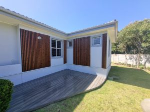 Utsnitt av hvit husfasade med to smale, vertikale trelamellpaneler fra WoodUpp, mørkbeiset og jevnt montert rundt vinduer, samt en grå, buet terrasse av WoodUpp terrassebord i forgrunnen. Bildet viser gressplen, lave busker og trær i hagen under klar blå himmel.