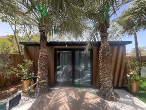 WoodUpp hageanneks med vertikale trelameller som fasadekledning, mørke metallprofiler og store glass skyvedører. Fronten prydes av to palmer, pottede planter og hengende lyspærer. Trelamellene er smale, naturfargete og jevnt montert i sort ramme på lys betongterrasse, i en solrik, privat hage.