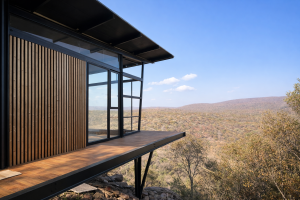 Et bilde av en moderne hytte med en terrasse som vender ut mot et vidstrakt landskap. Ytterveggen er kledd med WoodUpp Akustikkpanel, karakterisert av loddrette trespiler i en varm eikefarge. Disse panelene skaper en naturlig kontrast til de store glasspartiene og den sorte stålrammen på bygningen.