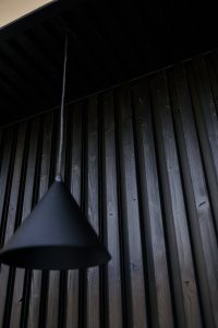 Bilde av WoodUpps mørkbehandlede, vertikale trepaneler på vegg og tak med tydelig trestruktur, årer, spor mellom bordene og synlige skruer; foran henger en svart, kjegleformet pendellampe i matt metall på tynn ledning, som skaper dype skygger og et moderne, minimalistisk uttrykk.