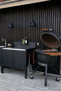 WoodUpp utekjøkken med svart skapmodul, rustfri vask og svart benkeplate står mot et sort vertikalt listpanel, med en liten hylle bak og to hengende sorte lamper. Ved siden av står en stor keramisk kamado-grill med trehåndtak og rulleunderstell. På benken en flaske olje og urtepotte.