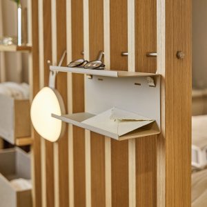 WoodUpp romdelersystem i lys eik med vertikale spiler, montert med to avtakbare beige metallhyller/oppbevaringsbrett, tynne stålholdere, små kroker og rund vegglampe med varmt lys. Øverste brett har briller, nederste en konvolutt; synlig messingbolt, naturlig trestruktur og diskret fargeskjema som gir praktisk oppbevaring.