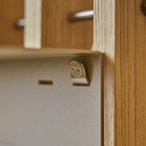 WoodUpp-produkt: nærbilde av en matt metallkrok festet til en tynn metallplate og montert på WoodUpp eikepanel. Kroken har et gravert sirkelmotiv (logo). Bildet viser detalj av materialer — børstet metall og varm eikefinér, med synlige metallbeslag i bakgrunnen og tydelig trestruktur.