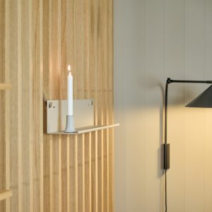 WoodUpp lamellvegg i lyst eikfiner med vertikale trelister, og en hvit WoodUpp vegghylle med integrert lysestake som holder et tent hvitt kronelys. Bildet viser tretekstur, hyllens matte overflate, minimalistisk design og en svart vegglampe i bakgrunnen som gir varmt, dempet lys og myk stemning i rommet.