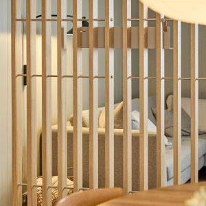 WoodUpp romdeler i lyst eik med jevne vertikale lameller, tynne metallstag og synlige festebraketter, brukt som romavskiller mellom spise- og stueområde. Bak skimtes en grå sofa med puter. Produktet er et modulært, monterbart WoodUpp lamell-system med kroker og festepunkter i varmt treverk med synlige årer.