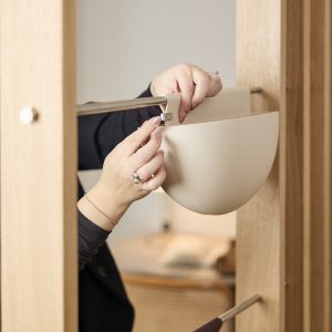 Nærbilde av hender som fester en beige WoodUpp hengende plantekrukke til en metallskinne på et lyst trepanel. Produktet viser en buet, matt plastpotte med integrert krok og et lite metallfeste som låser potten sikkert på skinnen. Stangen er børstet stål, treverket lyst eikefiner.