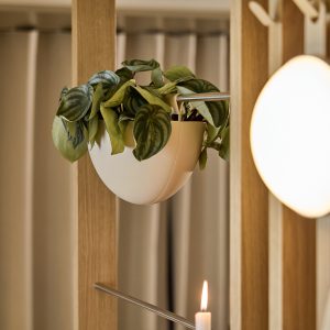 WoodUpp vises med en stilfull lys tre romdeler med vertikale lameller og horisontale metallstenger som holder en hvit, halvkul hengepotte fylt med grønne, hjerteformede blader. Bildet viser også et tent telys og en rund, varm lampe som gir myk stemningsbelysning.