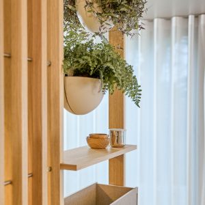 WoodUpp elegant romdeler i lyst tre med naturlig eikefinish, innebygd hylle, hengende hvite plantepotter festet i systemet og en romslig trekasse nederst. På hyllen ligger små treskåler og en blank metallkrukke, mens frodige grønne planter henger fra de innebygde ampelfestene.