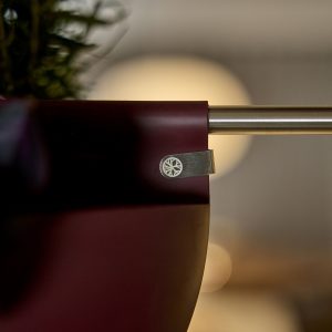 Nærbilde av en WoodUpp hengende plantepotte i burgunder, elegant festet til en glatt, rund metallstang med en liten, sølvfarget WoodUpp-klemme. Klemmen viser en innrisset sirkel med tre-hjul-logo og et dekorativt gravert motiv, og holder pottekanten godt fast. Myk, uskarp bakgrunn fremhever potte og logo.
