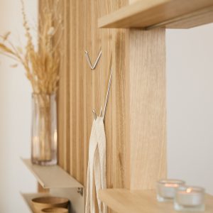 WoodUpp slatvegg i lyst eik med vertikale lameller, montert med blanke, V-formede kroker og tynne metallhyller, samt en åpen eikehylle over. Bildet viser en krok med hengende bomullsrem, små treboller på hylle, en glassvase med tørkede blomster og tente telys som dekor.