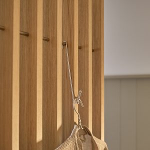 WoodUpp eikevegg med smale vertikale spiler og innebygde bronsefargede knagger, vist i nærbilde. Et langt metalloppheng fra WoodUpp henger i en av knaggene og holder en beige tekstilpose, med varm eikefarge og slanke, polerte detaljer.