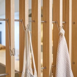 WoodUpp romdelere i lyst eiktre med slanke, horisontale rustfrie stålstenger og avtakbare WoodUpp-kroker i polert stål, brukt som oppheng for en stripete kjøkkenklut og en tung stoffpose. Nærbilde viser elegant, funksjonell og plassbesparende opphengsløsning, ideell for kjøkken, entré eller stue.
