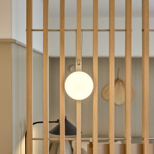 WoodUpp trelameller i lyst eik danner en slank romdelerskjerm med synlige metallstenger. Foran henger en rund, opalhvit WoodUpp-lampe festet med lærstropp og metallkrok. Bildet viser moderne, skandinavisk interiør med mykt, varmt lys og naturlig trestruktur, delikat, minimalistisk og funksjonell estetikk.