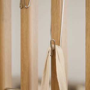 Detaljfoto av WoodUpps lyse eikelameller i et romdelersystem: vertikale, finerte trestaver med tydelig åretegning og glatt, matt overflate, hvor slanke, forkromede og U-formede metallkroker er montert i spor. En kremfarget, vevd skulderrem henger i en krok, og bildet fremhever trestruktur og minimalistisk design.