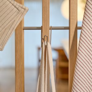 WoodUpp eikepanel med horisontal stålstang og to enkle stålkroker; en beige bomullsrem henger midt på, og stripete og rutete kjøkkenhåndklær henger til høyre og venstre. Nærbilde viser treets varme årringer, metallglans og tekstilfibre i naturlige toner, med enkel, minimalistisk finish som fremhever nordisk design.