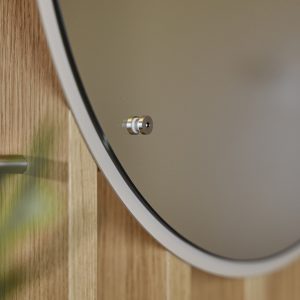 Nærbilde av WoodUpp rundt speil med hvit ramme, festet med et lite metallavstandsstykke, montert på lyst eike trepanel fra WoodUpp. Del av grøntmetall krok og uskarp plante til venstre gir kontekst, fokus på speilets feste og trepanelenes vertikale detaljer.