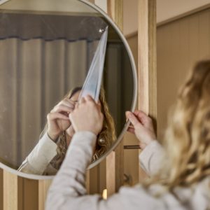 Person fjerner beskyttelsesfilm fra et rundt WoodUpp-speil med hvit ramme, montert på WoodUpp lyst trepanel med vertikale lameller. Nærbilde viser speilets refleksjon, trestruktur, hendene som drar av plasten og en delvis synlig person med langt, bølget hår i varmt interiør.