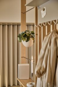 WoodUpp romdeler/garderobe i eik med tynne vertikale sprosser, hylle og metallstang med knagger. På hyllen en tent lysestake og bøker, og en hvit hengende plantekule festet i stangen. En beige kåpe henger på knaggene, og en marmorring som dekorasjon i dempet, varmt lys.