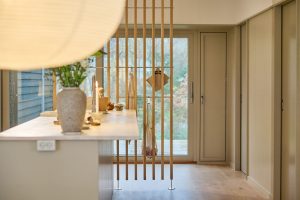 WoodUpp spilevegg i lys eik deler kjøkkenet fra terrassen; vertikale, slanke trefinérspiler med diskrete metallfester fungerer som romdeler og oppheng for kluter og nett. Bildet viser også en kjøkkenøy med vase, tent lys og nøytrale materialer. Spilene har en slank profil og naturlig, oljet overflate.