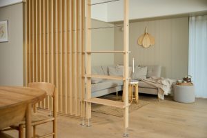 WoodUpp romdeler i lyst eik med tynne, jevnt plasserte vertikale lameller som deler spisestue og stue, kombinert med en WoodUpp åpen hylleseksjon/stige i eik med tynne stålstag, lave hyller og justerbare metallføtter. Naturlig eikefinish og synlige metallfester gir et minimalistisk, skandinavisk uttrykk.