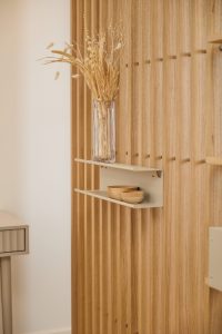 WoodUpp slatvegg i lyst eiktre med vertikale lameller, montert med to beige, flytende metalhyller fra WoodUpp med diskrete beslag. Øverste hylle har en høy glassvase med tørkede strå; nederste hylle holder to små elegante treskåler på den varme, teksturerte treoverflaten