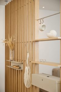 WoodUpp romdel i lyst eikfiner med vertikale lameller, innebygde hyller, metallkroker og beige oppbevaringsboks. Bildet viser WoodUpp-hyller med liten keramikk-kopp på hengerekke, nettingpose, glassvase med tørkede strå, små trefat, to telys og en hvit skjellfigur i elegant, minimalistisk, skandinavisk stil.