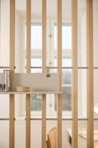 WoodUpp romdeler i lyst eiketre med tynne vertikale spiler og metallstag, med en avtakbar grå hylleplate fra WoodUpp festet med liten magnetkrok. På hyllen står en liten rund trebolle og en blank metallkanne, i et lyst, minimalistisk rom og kontorområde.