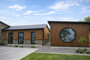 Moderne eksteriør av to bygninger kledd med WoodUpp sin fasadekledning. De stående trespilene har en varm, gyllenbrun farge som står i kontrast til den mørke steinen nederst og det svarte metalltaket. Bygget til høyre har et markant stort, rundt vindu, mens det venstre har høye, smale vinduer.