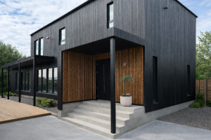 Bildet viser et moderne hus med mørk, stående kledning. Det overbygde inngangspartiet er fremhevet med WoodUpp akustikkpaneler. Disse vertikale spileveggene i varmt treverk gir en lun kontrast til den svarte fasaden, med en bred betongtrapp og en stor potteplante foran inngangsdøren.